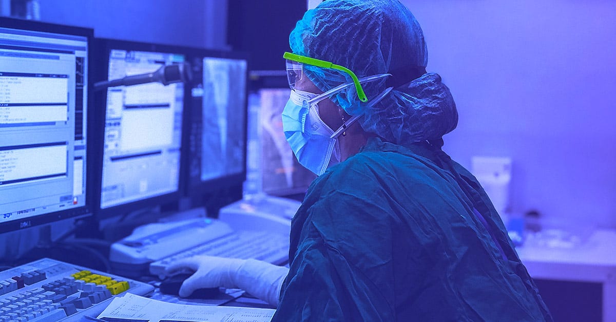 Healthcare worker in full scrubs, cap, and mask using a computer